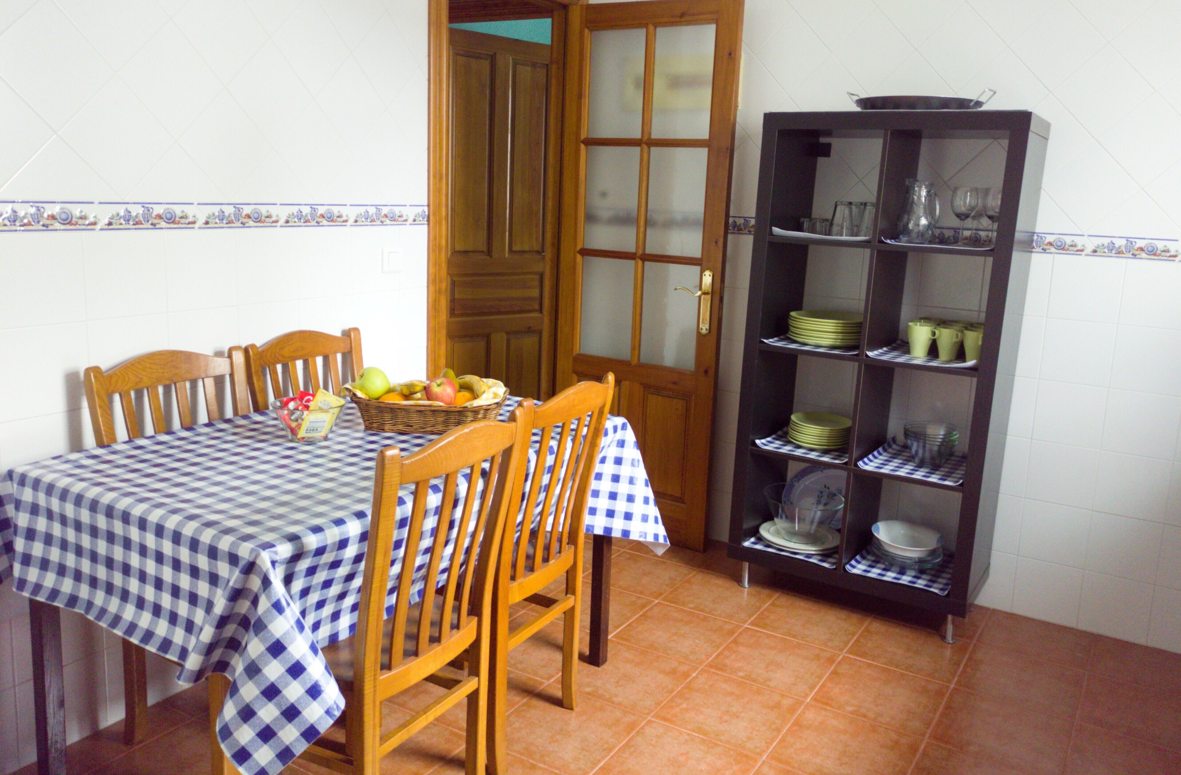 A dining area with a checkered tablecloth, wooden chairs, a fruit basket, and a glass cabinet with dishes.