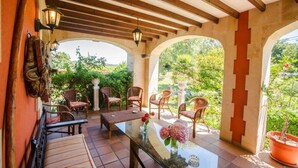 A covered outdoor seating area with wicker furniture, a glass table, and potted plants. - Posada Río Cubas (Ribamontan al Monte)