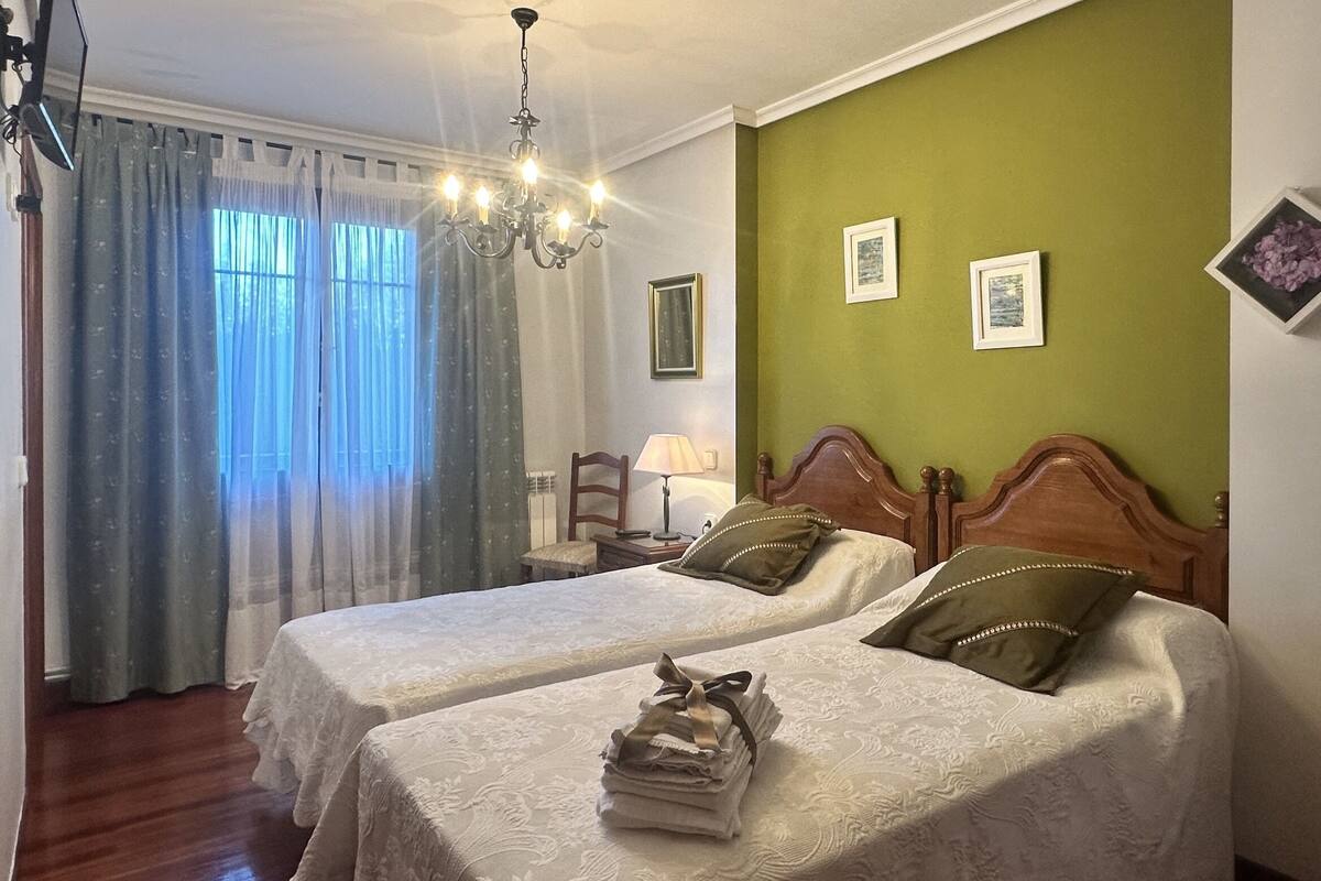 A hotel room with two beds, a chandelier, a nightstand with a lamp, and a window with curtains.