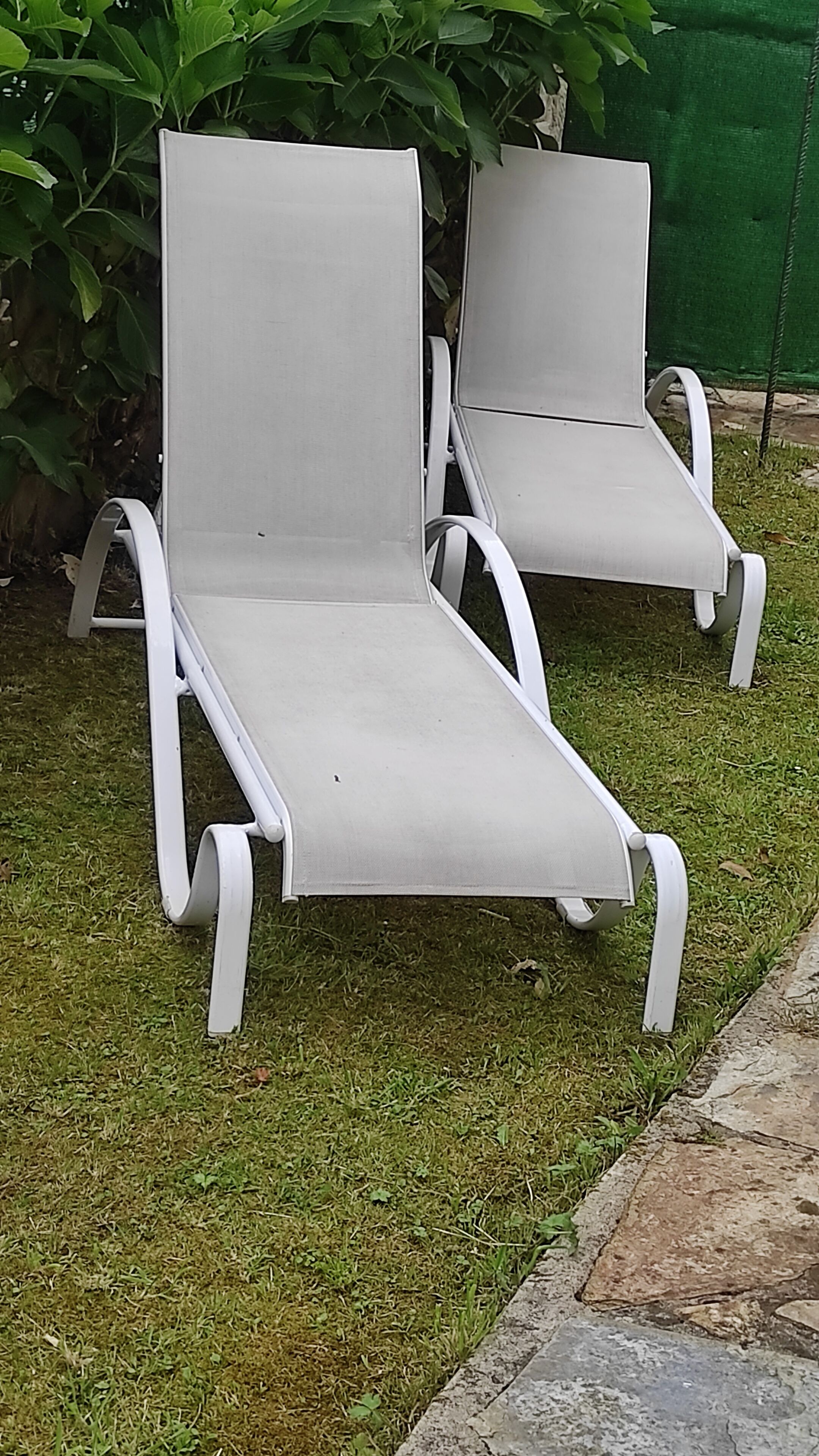 Two white lounge chairs with metal frames, positioned on a grassy area with a stone border.