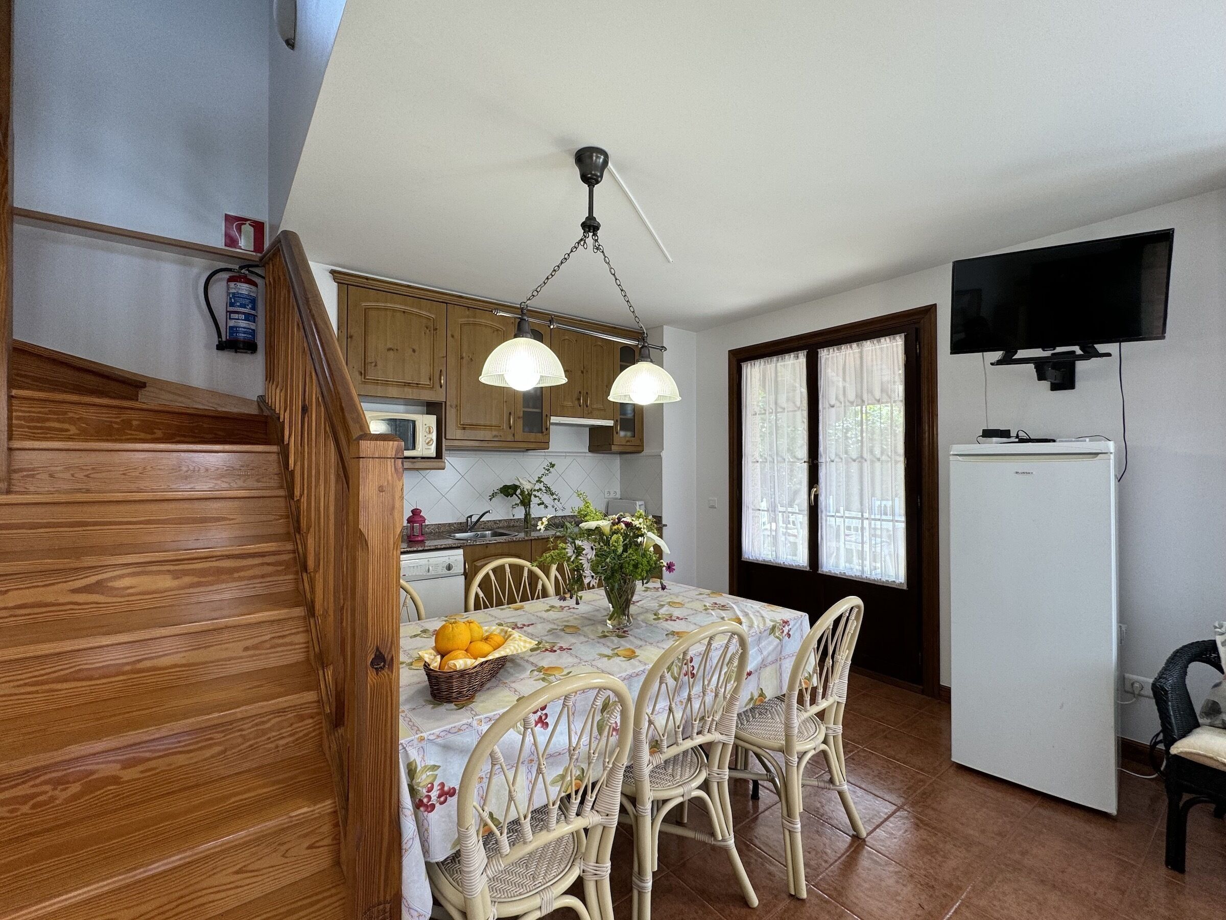 A kitchen with wooden cabinets, a dining table with chairs, a refrigerator, and a television mounted on the wall.