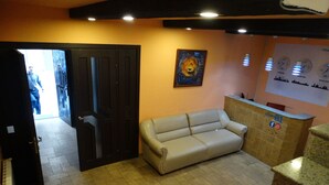 Un área de recepción con un sofá, una puerta que da al exterior, un cuadro en la pared y un mostrador con un cartel.