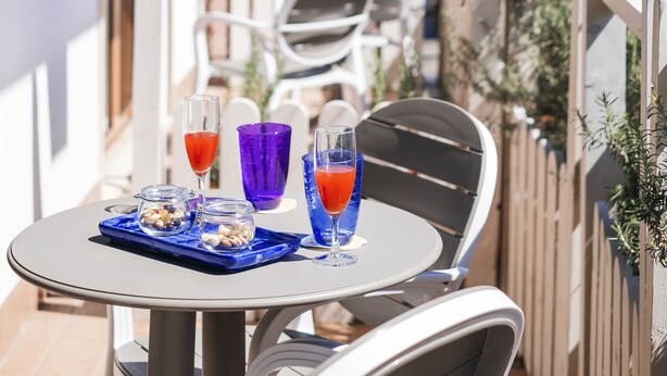 A balcony with a table set for two, featuring a glass of red drink and a glass of blue drink, and a small tray with nuts and a bottle.