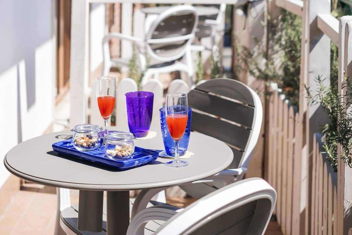 A balcony with a table set for two, featuring a glass of red drink and a glass of blue drink, and a small tray with nuts and a bottle.