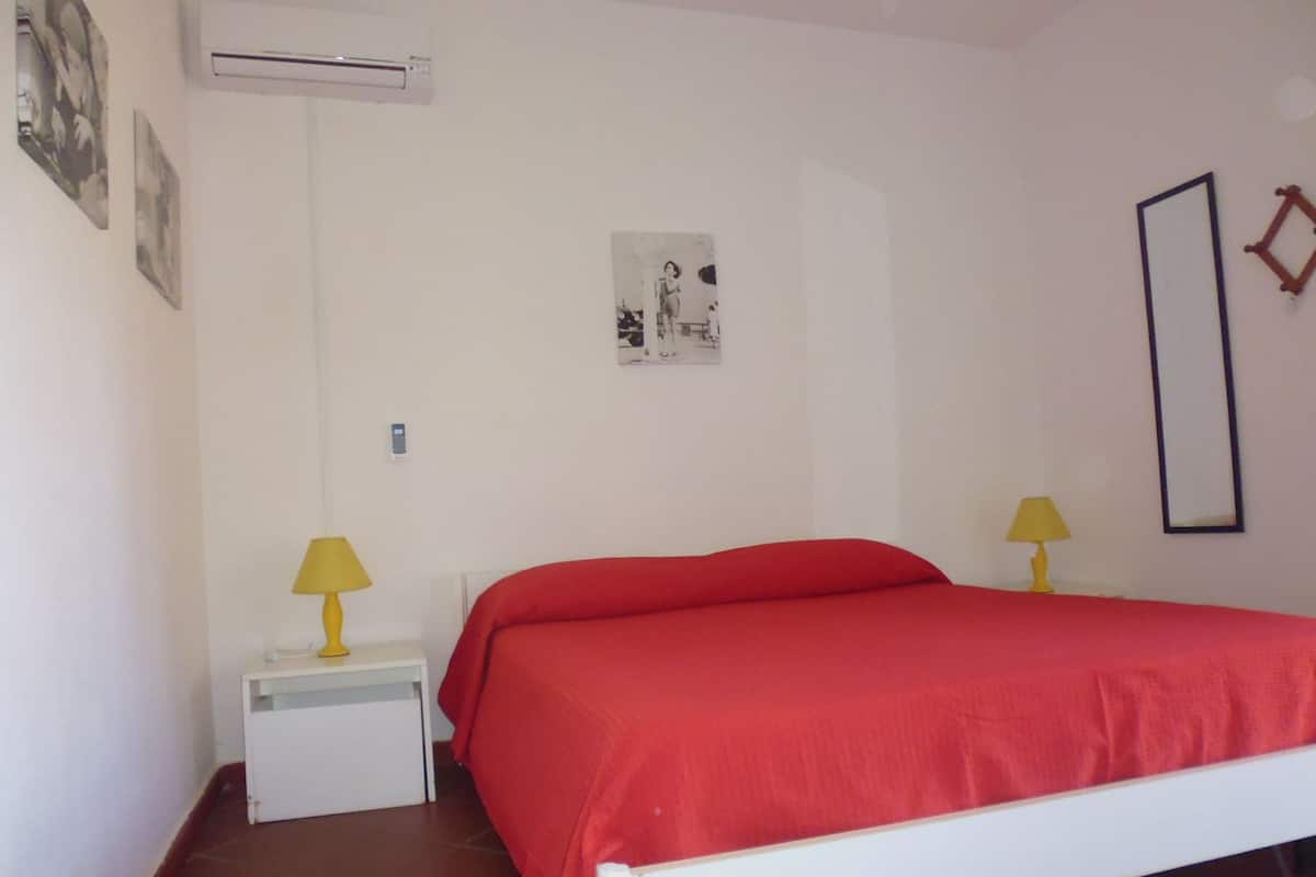 Une chambre avec un lit blanc, une literie rouge, deux lampes de chevet, une petite table de chevet, un appareil de climatisation et une affiche accrochée au mur.
