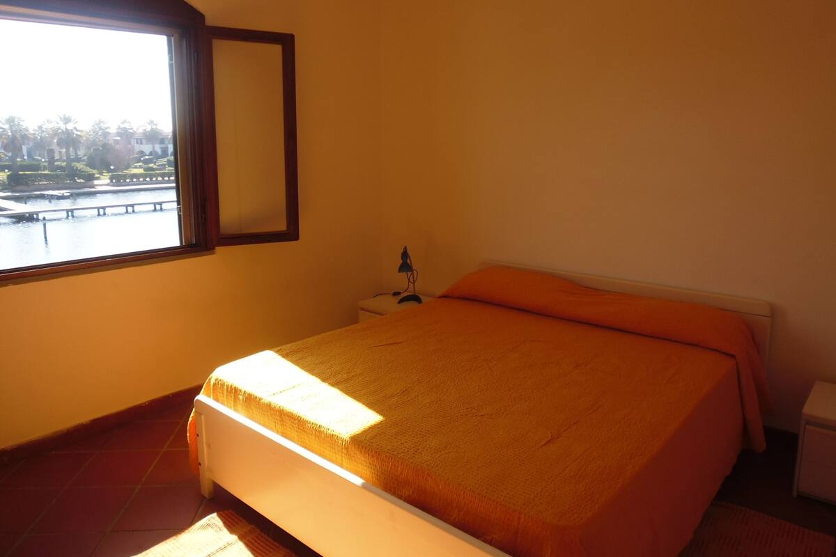 Une chambre à coucher comprenant un lit, une table de chevet, une lampe et une fenêtre donnant sur un parc et un plan d’eau.