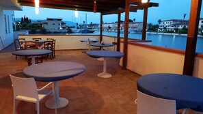 An outdoor terrace with round tables and chairs, overlooking a marina with boats and buildings in the background. - Residence La Bricola Laghi di Sibari (Cassano allo Ionio)