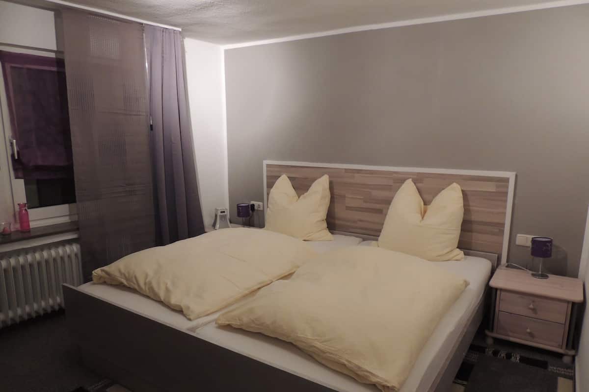 A neatly made bed with two pillows, a bedside table with a lamp, and a window with curtains.
