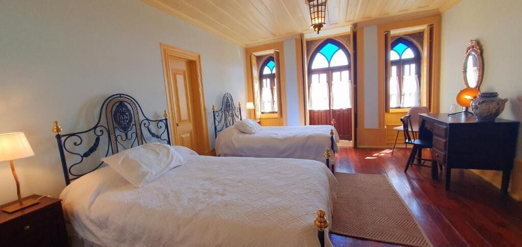 Habitación de hotel con dos camas, un escritorio con lámpara y una silla. Se observan ventanas con vidrieras.