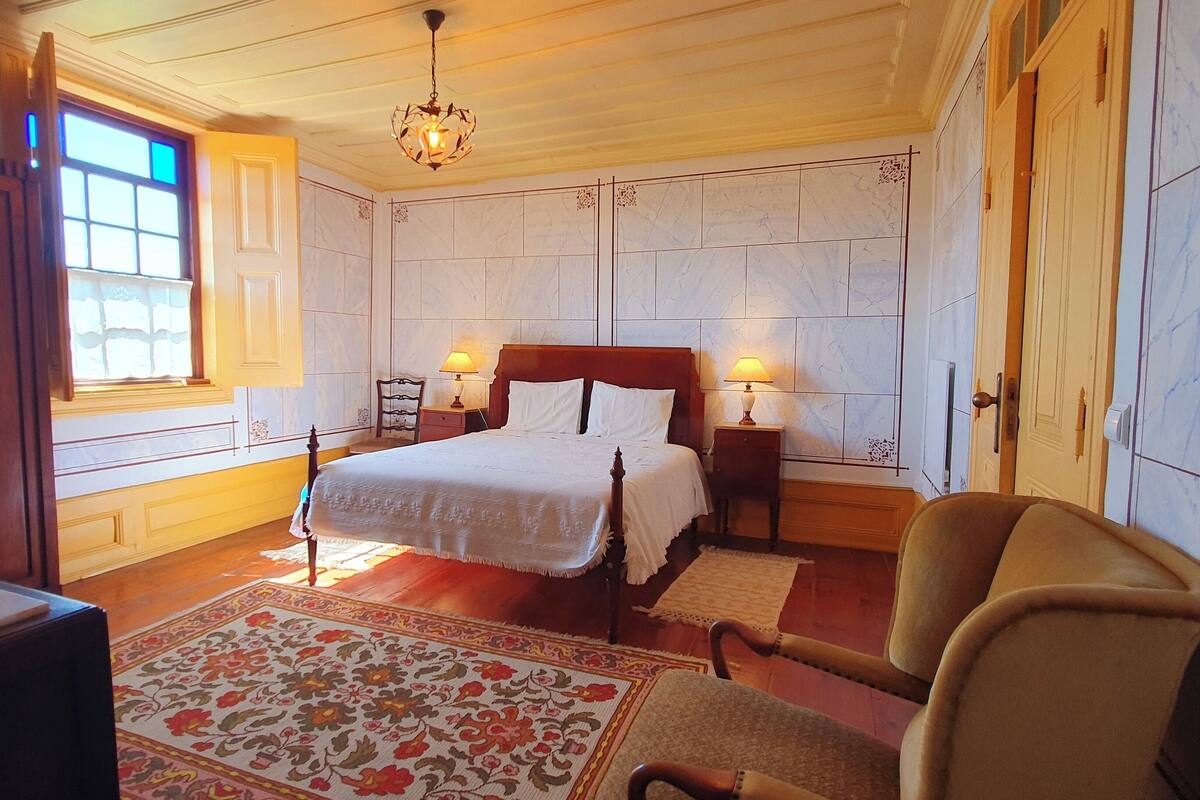 A bedroom with a large bed, two bedside tables with lamps, a wooden chair, a decorative fan, and a patterned rug.
