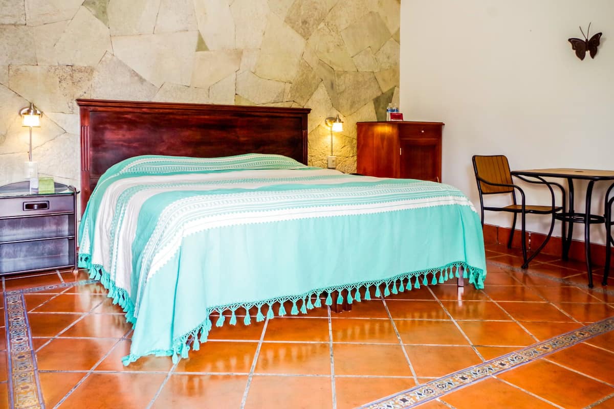 A bedroom with a large bed, a nightstand with a lamp, a wooden dresser, and a small table with chairs.