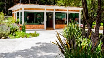 Un pequeño edificio de ladrillo con amplios ventanales, rodeado de vegetación y un camino pavimentado.