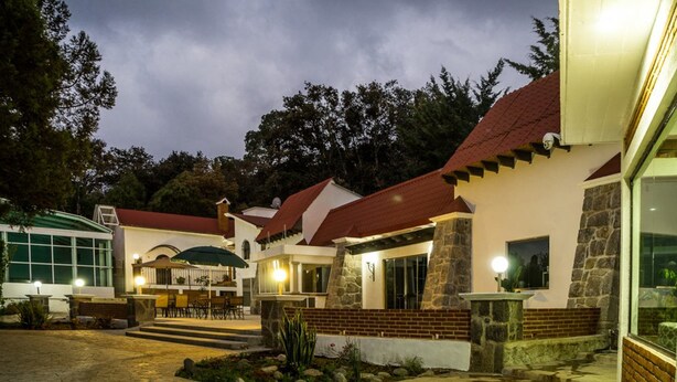 Un patio bien iluminado con un sendero de piedra, un área de comedor con mesas al aire libre y un edificio con techo rojo y muros de piedra.