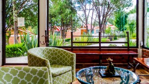 Habitación con un ventanal amplio que da a un jardín y edificios. Hay dos sillas estampadas en verde y una pequeña mesa con una estatua.