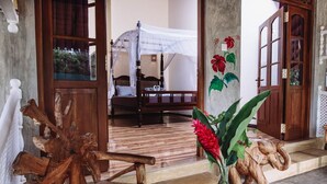 A room with a bed, a wooden chair, a table with a vase, and a door with a floral painting. - Thambili cabanas (Hikkaduwa)