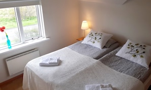 Un dormitorio con dos camas, una ventana que da a zonas verdes, una mesita de noche con lámpara y un radiador.