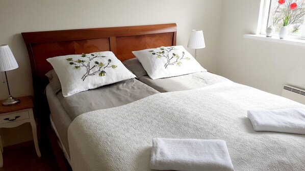 Una cama bien hecha con dos almohadas, una lámpara de noche y una ventana con flores.