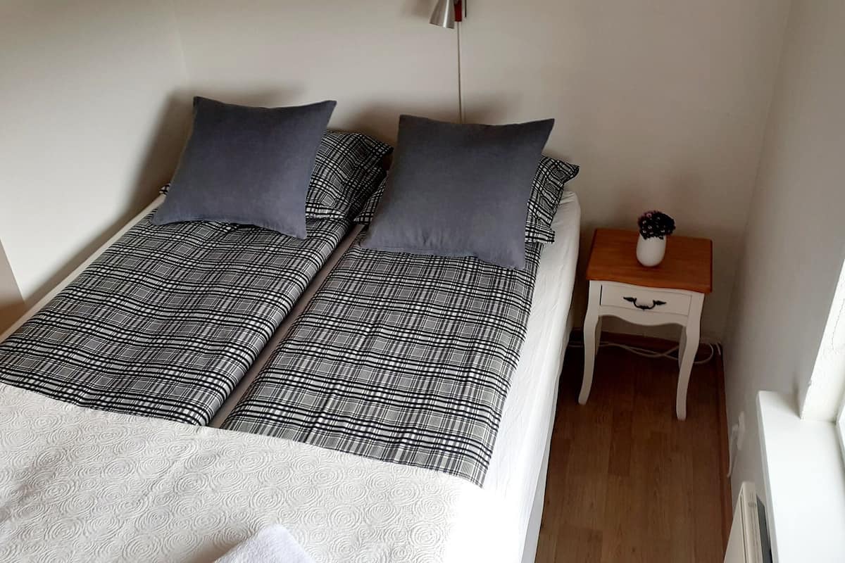 A single bed with a black and white checkered bedspread and two pillows. There is a small wooden nightstand with a vase of flowers beside the bed.