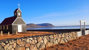 A small church with a red roof and a cross on top, surrounded by a stone wall and a grassy area.