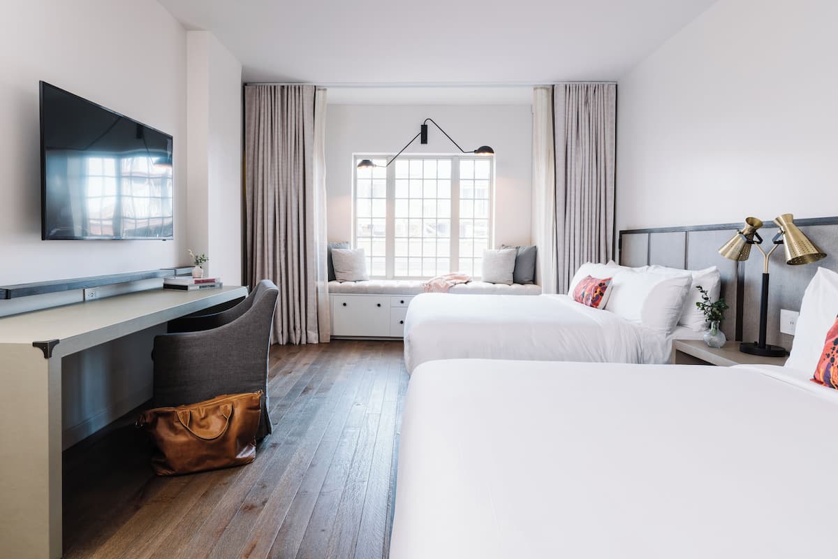 Ein Hotelzimmer mit großem Flachbildfernseher, Schreibtisch mit Stuhl, zwei Betten, einem Fenster mit Vorhängen und einem Nachttisch mit Lampen.