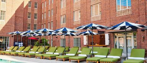 A poolside area with green lounge chairs and umbrellas, a brick building in the background, and a clear sky.