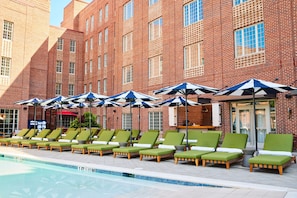 A poolside area with green lounge chairs and umbrellas, a brick building in the background, and a clear sky.