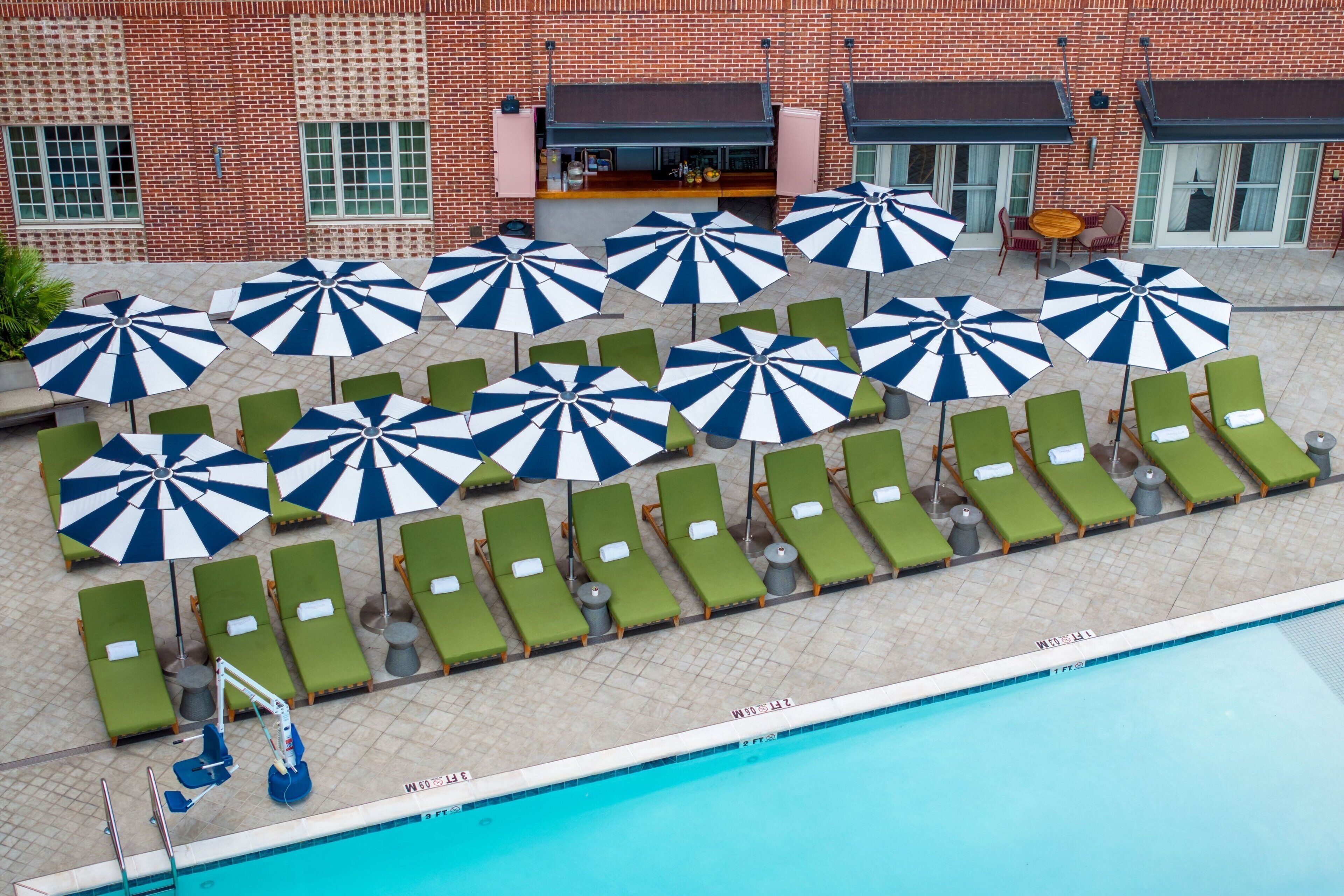 A poolside area with multiple lounge chairs and umbrellas, a brick building in the background, and a clear blue pool.