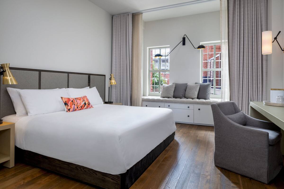 Ein modernes Hotelzimmer mit einem großen Bett, einem Schreibtisch und Blick auf ein Ziegelgebäude durch ein Fenster.