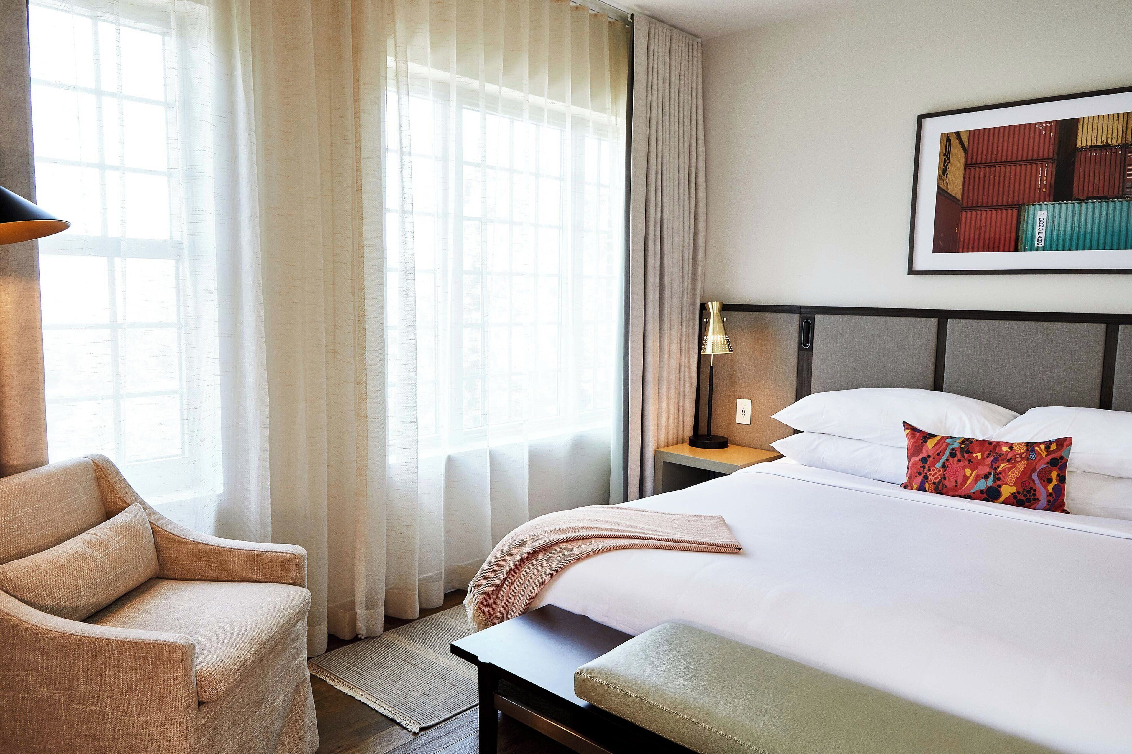 A hotel room with a bed, a chair, a nightstand, and a window with curtains.