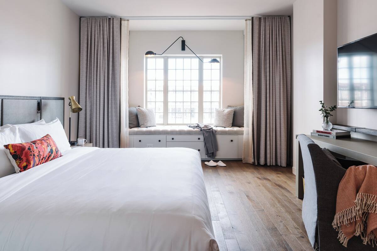 Ein modernes Hotelzimmer mit einem großen Bett, einem Schreibtisch mit Flachbildfernseher, einer Sitzbank unter einem Fenster mit Vorhängen und Blick auf die Stadt.