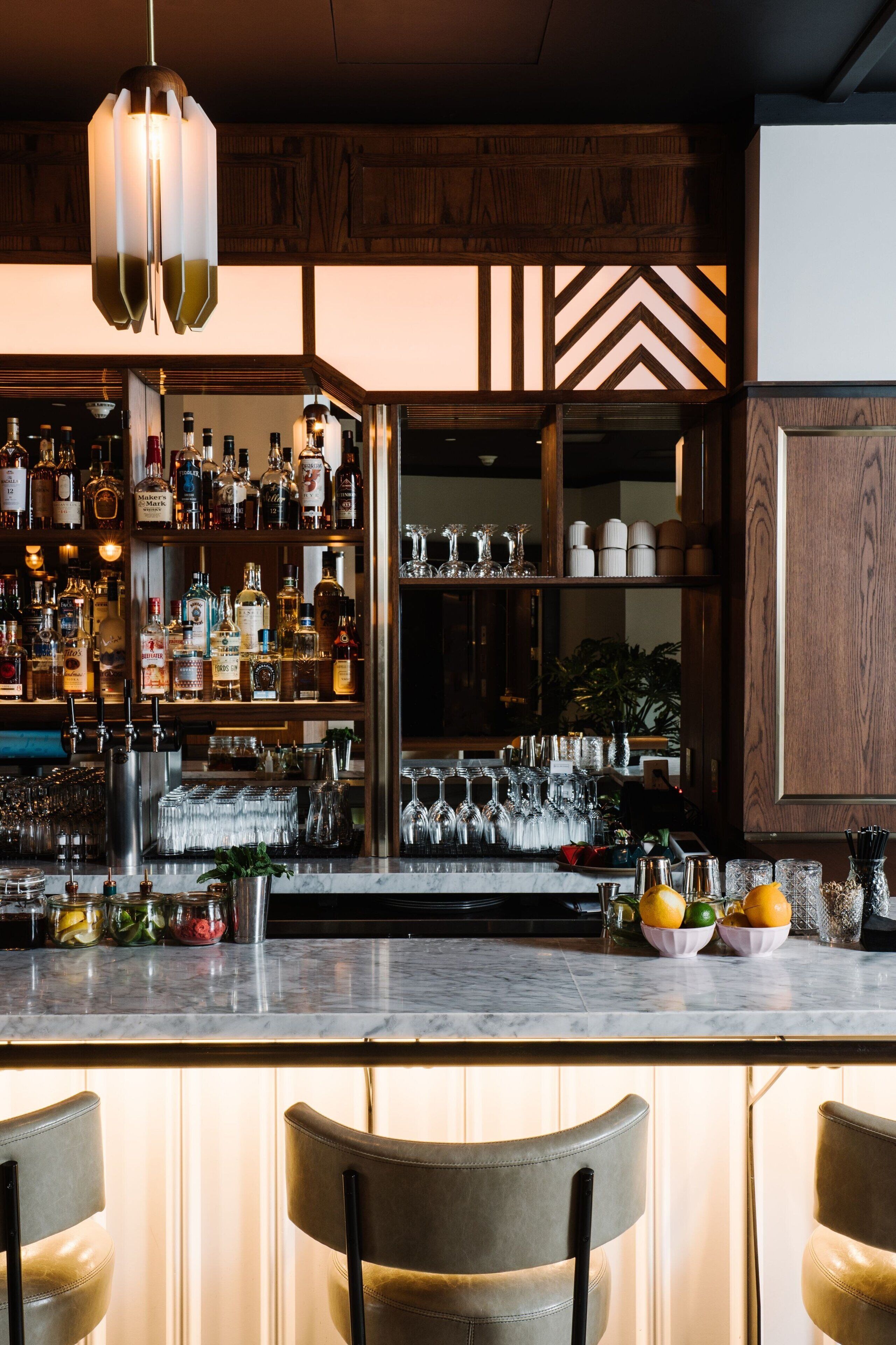 A bar area with a wooden bar counter, shelves of bottles, glasses, and a hanging light fixture.