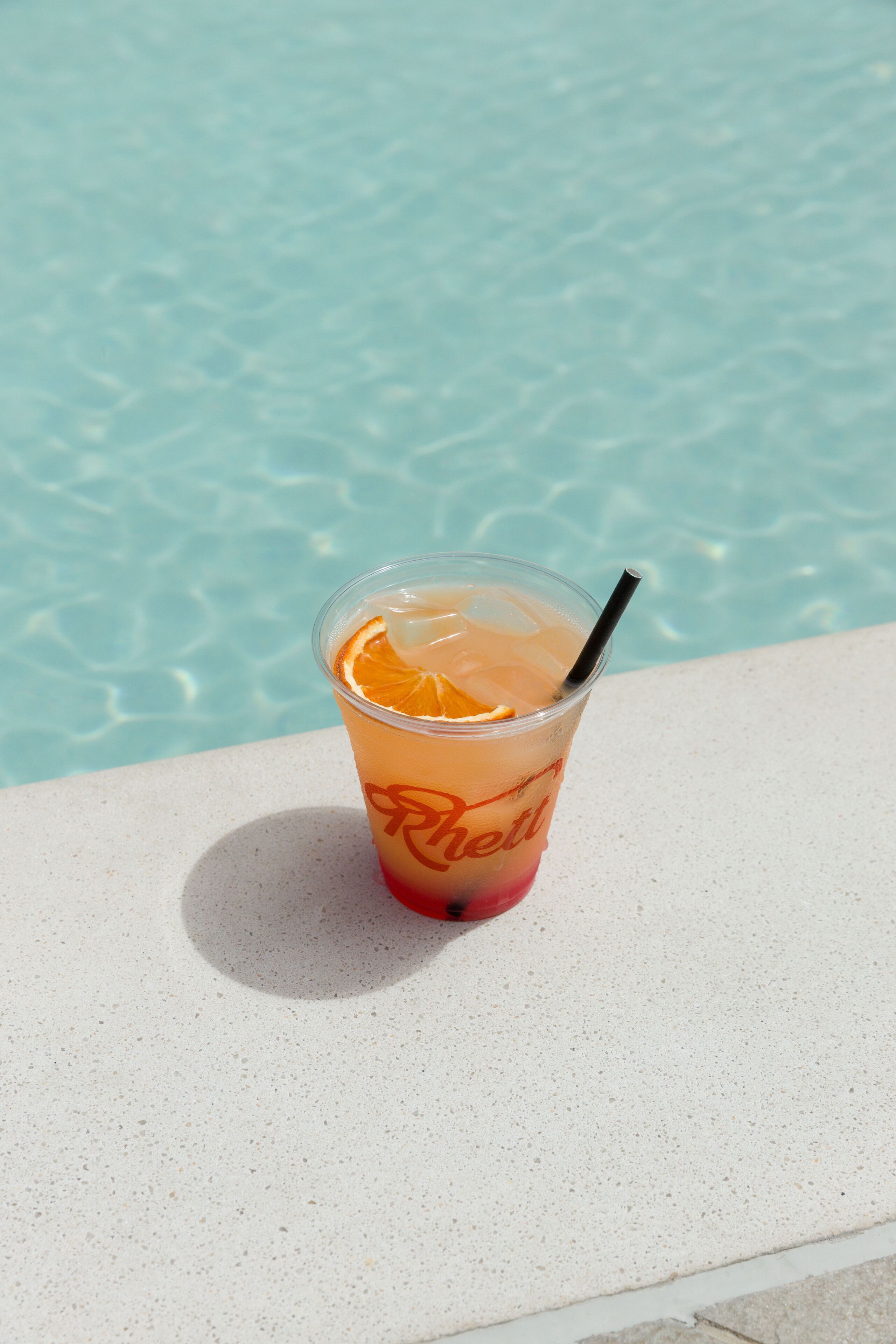 A clear plastic cup with a straw and an orange slice, placed on a white surface near a pool.