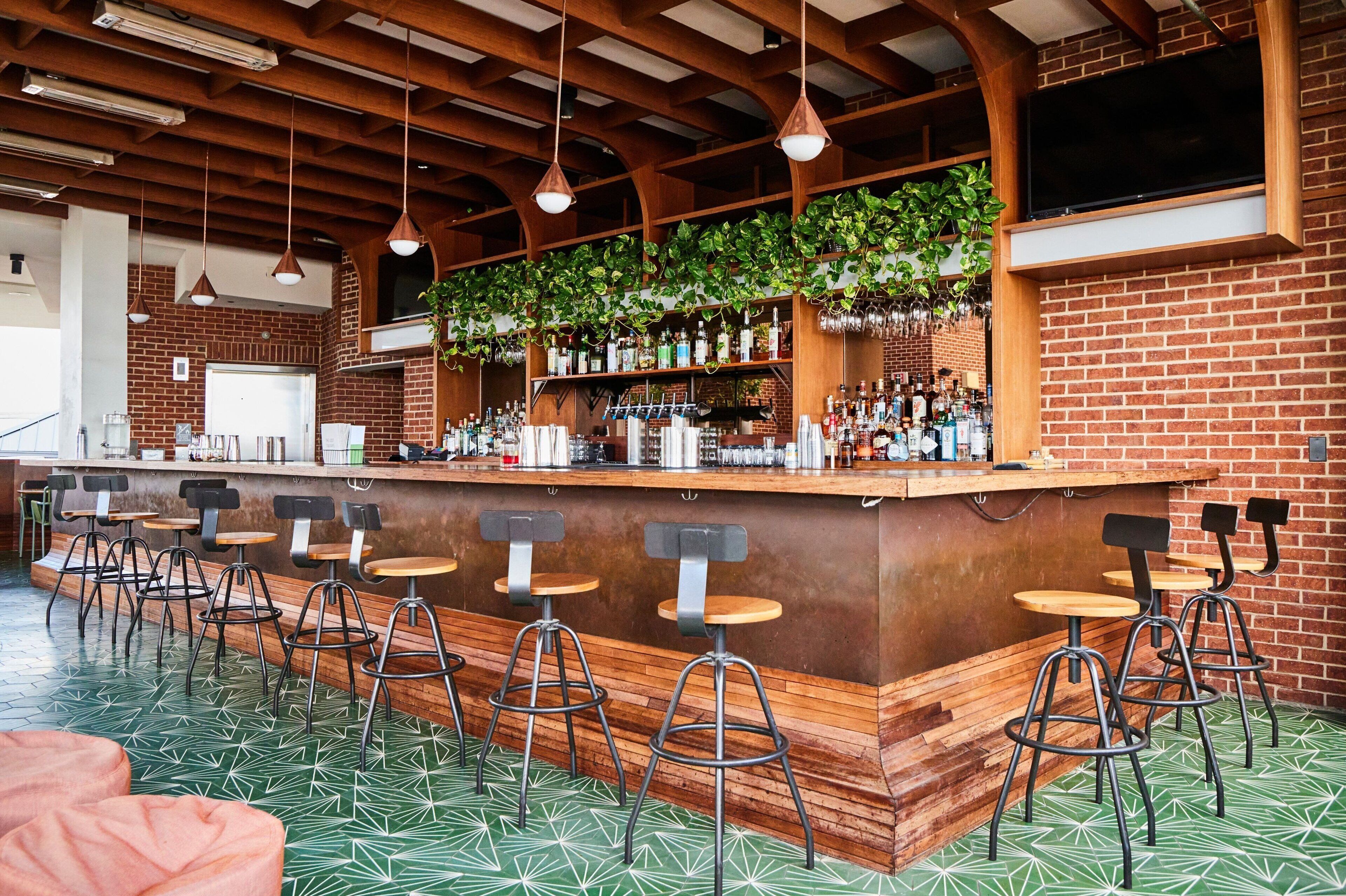 Eine Bar mit Backsteinwand, Holzbalkendecke, hängenden Pflanzen und Barhocker.