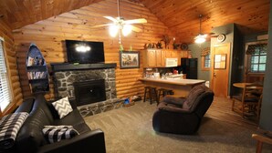 L’intérieur d’un chalet confortable, avec une cheminée en pierre, un téléviseur fixé au-dessus, un canapé confortable, un coin repas avec un bar et une cuisine dotée de meubles en bois.