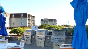 Outdoor seating with white chairs and blue umbrellas, a barbecue grill, and a stone wall. - 2/2 Ocean View Condo at APATB Resort (Atlantic Beach, NC)