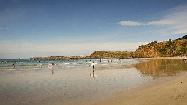 A beach with people walking and surfing, calm water, and a clear sky.