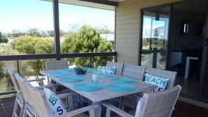 A wooden deck with a table set for four, surrounded by wicker chairs with 'BEACH' pillows.