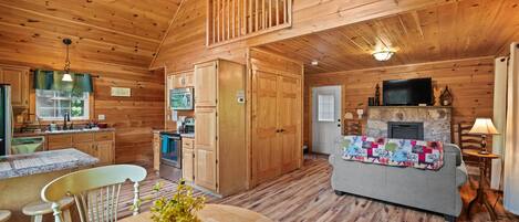 A wooden cabin interior with a dining area, kitchen, and living room featuring a fireplace.
