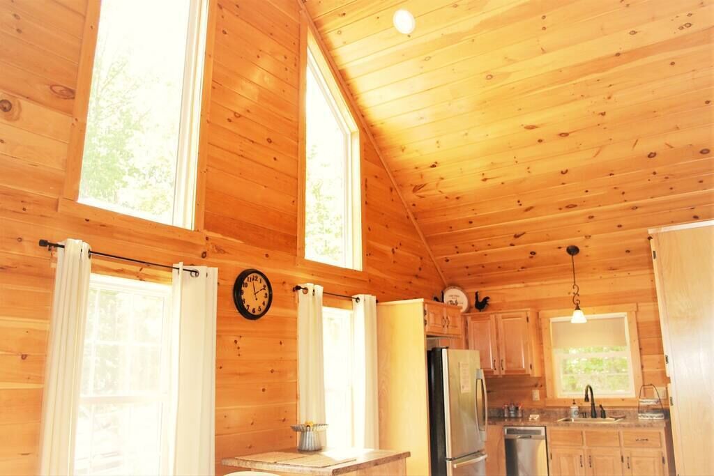A wooden cabin interior with a kitchen, large windows, and a clock on the wall.