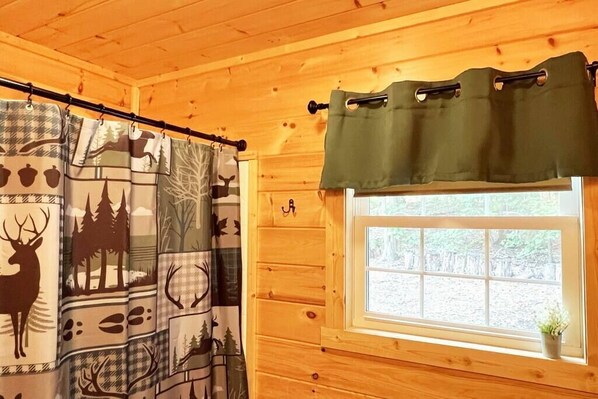 A cabin interior with a window, wooden walls, a curtain with deer and forest patterns, and a green curtain above the window. - The White Pine Loft: Perfect for Couples! (Stanley)