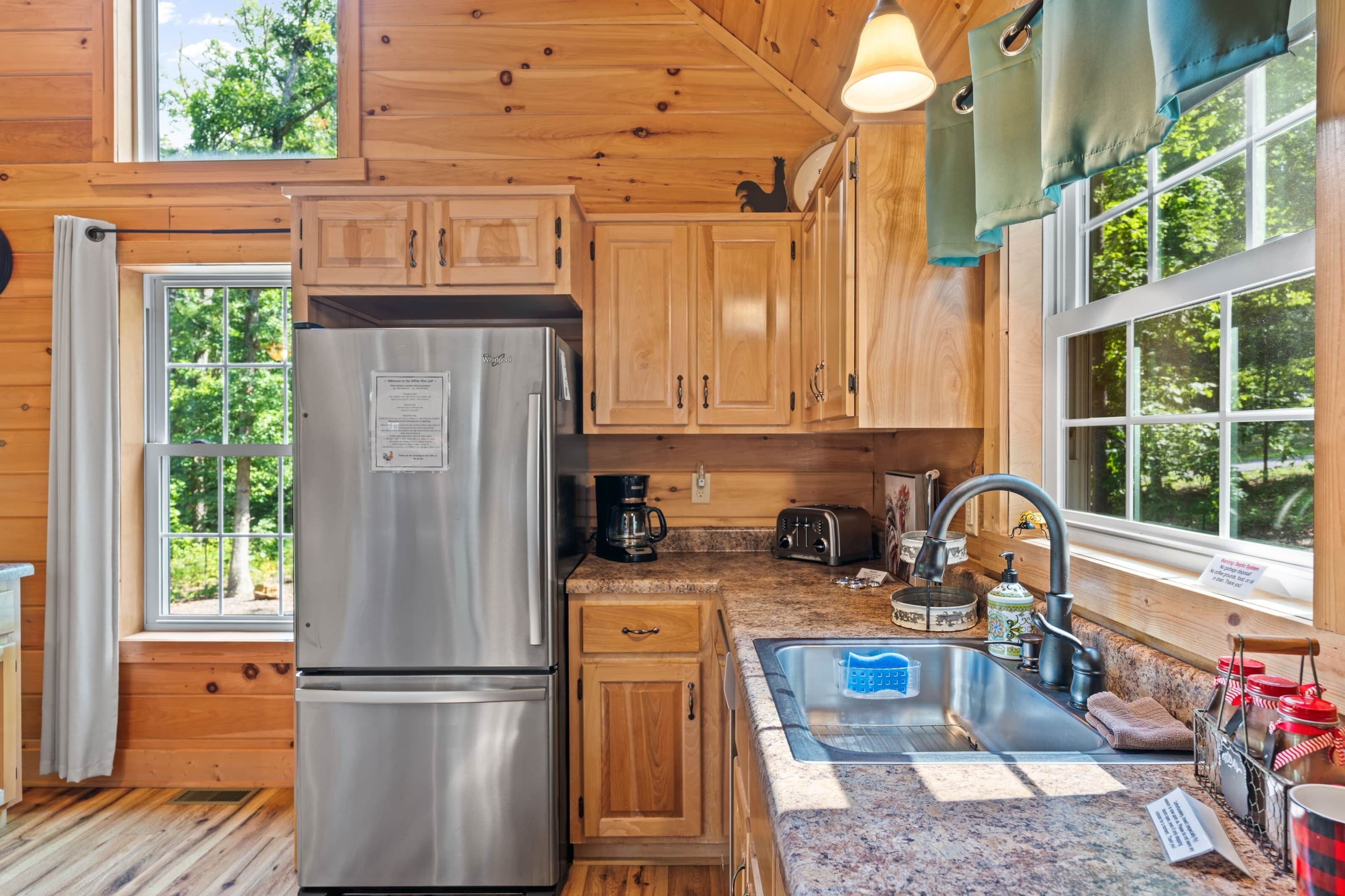 A wooden cabin kitchen with a stainless steel refrigerator, wooden cabinets, a granite countertop, a sink, and a window with a view of trees.