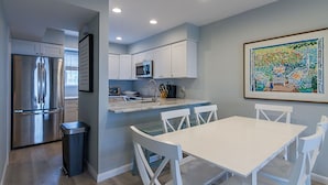 A modern kitchen with white cabinets, a stainless steel refrigerator, and a dining area with a white table and chairs. - Ocean View and only steps to the sand !! (Fenwick Island)
