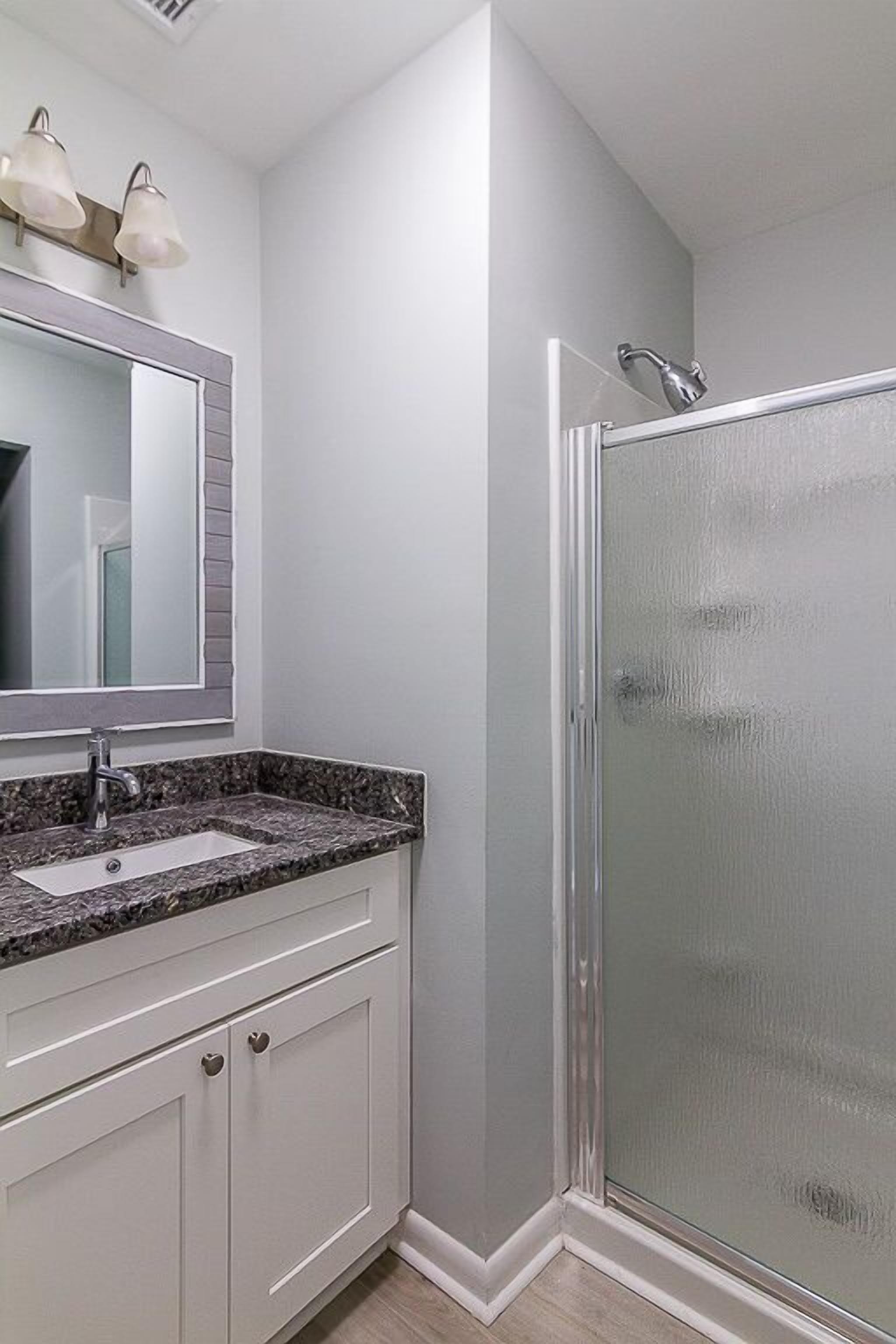 A bathroom with a granite countertop sink, a large mirror, frosted glass shower enclosure, and white cabinetry.