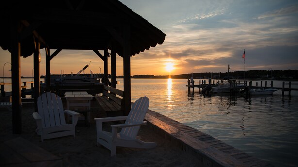 A waterfront scene with a wooden dock, two white Adirondack chairs, and a covered seating area. Boats are docked at the pier, and the sun is setting over the water. - Middle of Dewey ,On the Bay, Close to the Beach  (Dewey Beach)