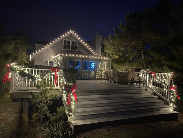 A house with a deck decorated with lights and garlands, a covered outdoor seating area, and a Christmas tree.