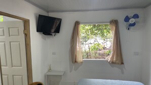 Una habitación con una cama, un televisor montado en la pared, una ventana con cortinas, una mesita y un ventilador.