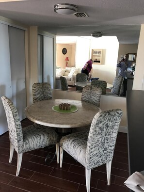 Un coin repas avec une table ronde et des chaises à motifs, un canapé en arrière-plan et une personne portant un t-shirt violet, penchée en avant.