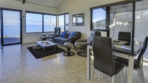 A spacious living room with a large window offering a view of the sea, a comfortable seating area with a sofa and chairs, and a glass table with chairs. - Park Beach Retreat - Ideal for couples (Dodges Ferry)