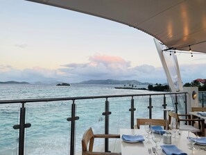 A dining area with tables set for two, overlooking a body of water with distant land and islands. - BEST VALUE! New reno, Beach 10' from door, water 50' away WE MOVED ID 5216594 !! (Red Hook)