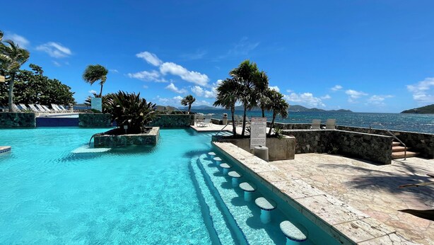 A clear blue pool with steps leading into the water, surrounded by lounge chairs and palm trees, with a view of the ocean and distant hills. - BEST VALUE! New reno, Beach 10' from door, water 50' away WE MOVED ID 5216594 !! (Red Hook)